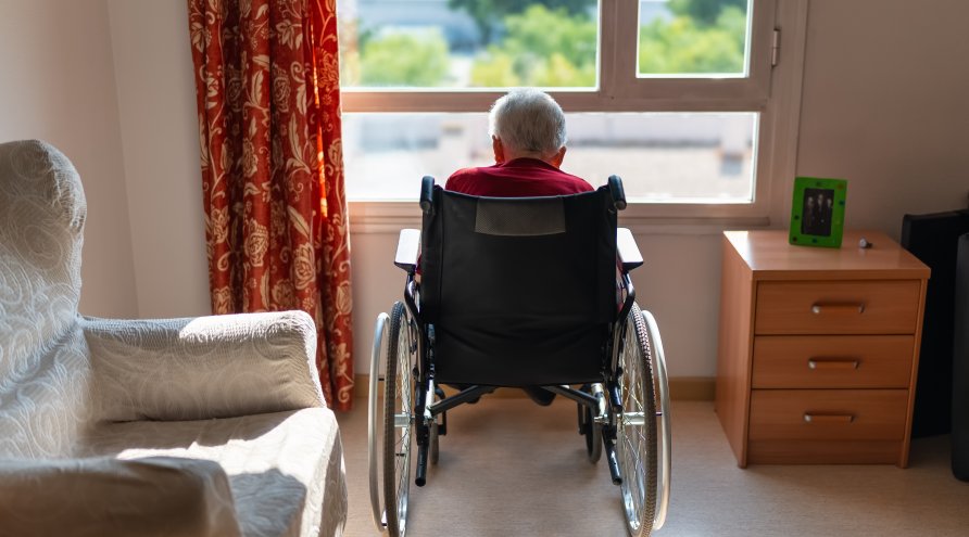 En gammel mann sitter alene i en rullestol og ser ut av vinduet