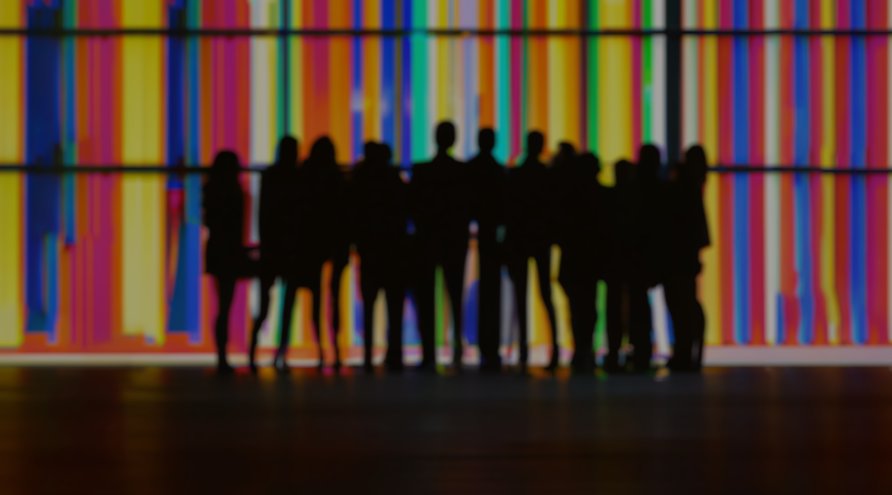 Gruppe mennesker, konturer i svart som er bluret, foran en vegg med striper i regnbuefarger.