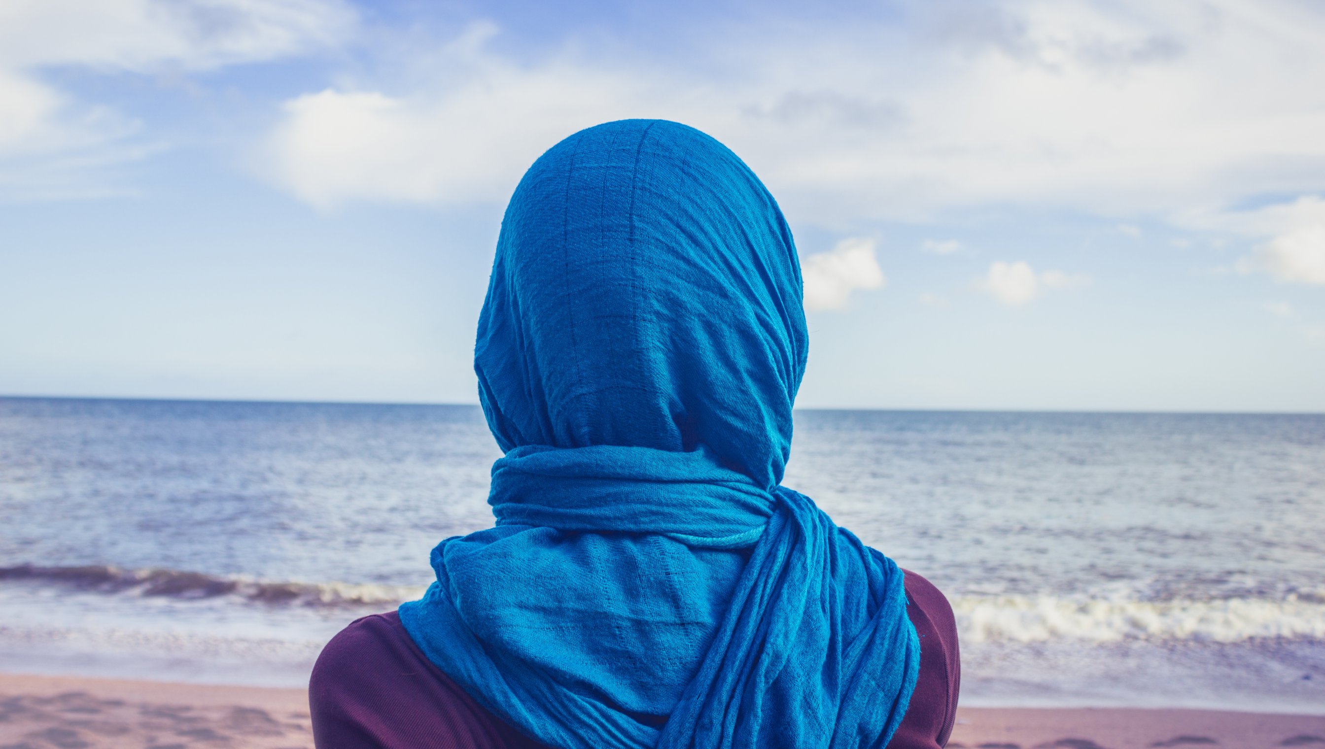 Kvinne med blå slør på hode, som står på en strand å ser ut over havet