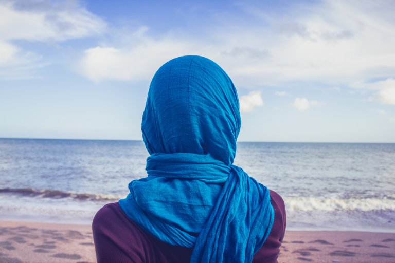 Kvinne med blå slør på hode, som står på en strand å ser ut over havet