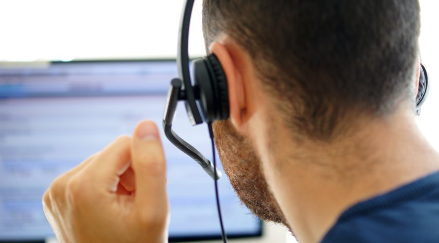 Mannlig bakfra sitter i telefon med headsett og kikke på dataskjerm.