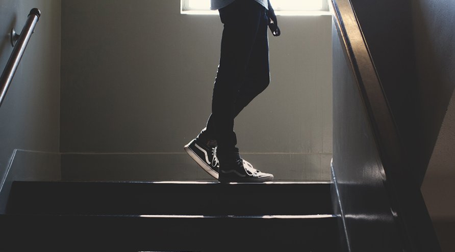 En ung person går oppover i en mørk trappeoppgang kledd i sneakers og bukse, bare nedre del av skjorten vises. Det kommer litt dagslys inn fra vinduet overst på bildet. 