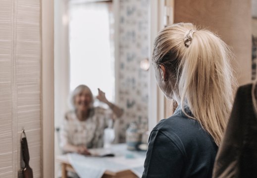 En kvinne fra hjemmetjenesten er i gangen til eldre kvinne som sitter ved kjøkkenbordet med en arm hevet. 