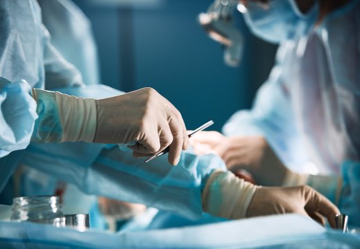 Hånd holder en skalpell under en operasjon. Helsepersonell kledd i sterile klær, med plasthansker og munnbind. Foto.