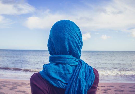 Kvinne med blå slør på hode, som står på en strand å ser ut over havet