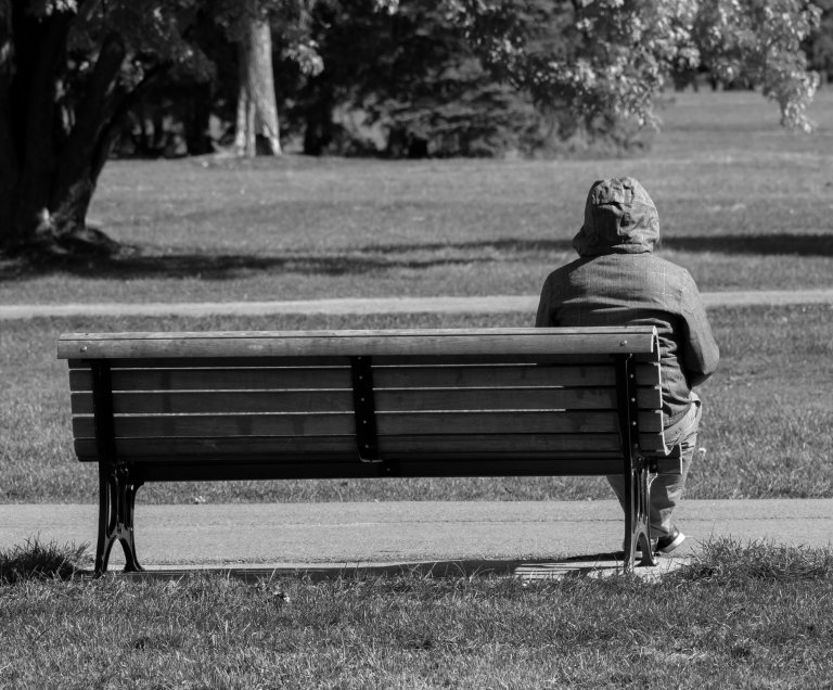 Illustrasjonsfoto av en person med ryggen til, som sitter alene på en benk i en park, i gråtoner.