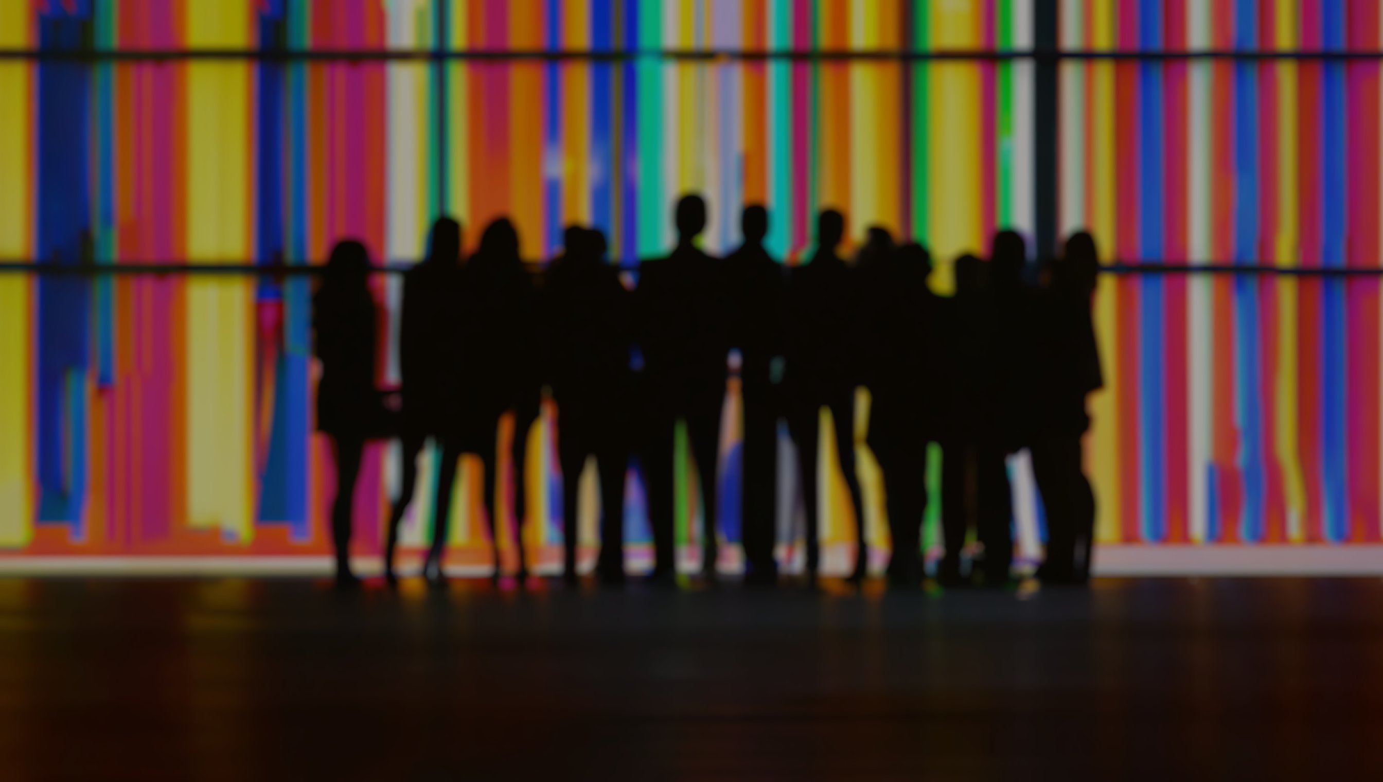 Gruppe mennesker, konturer i svart som er bluret, foran en vegg med striper i regnbuefarger.