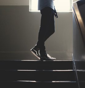 En ung person går oppover i en mørk trappeoppgang kledd i sneakers og bukse, bare nedre del av skjorten vises. Det kommer litt dagslys inn fra vinduet overst på bildet. 