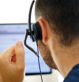Mannlig bakfra sitter i telefon med headsett og kikke på dataskjerm.