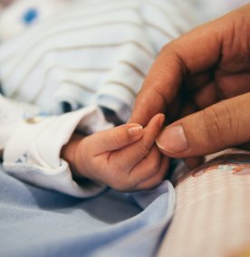 Babyhånd på et blått teppe, tre fingre berøres av en hånd fra en voksen person.